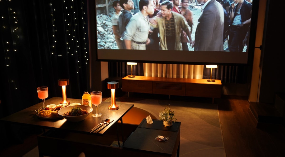A cozy home cinema setup is featured, showcasing a large screen displaying a movie scene. A low table accompanies bright drink glasses and a bowl of snacks. Soft lighting surrounds the space, enhancing the atmosphere for an inviting viewing experience.