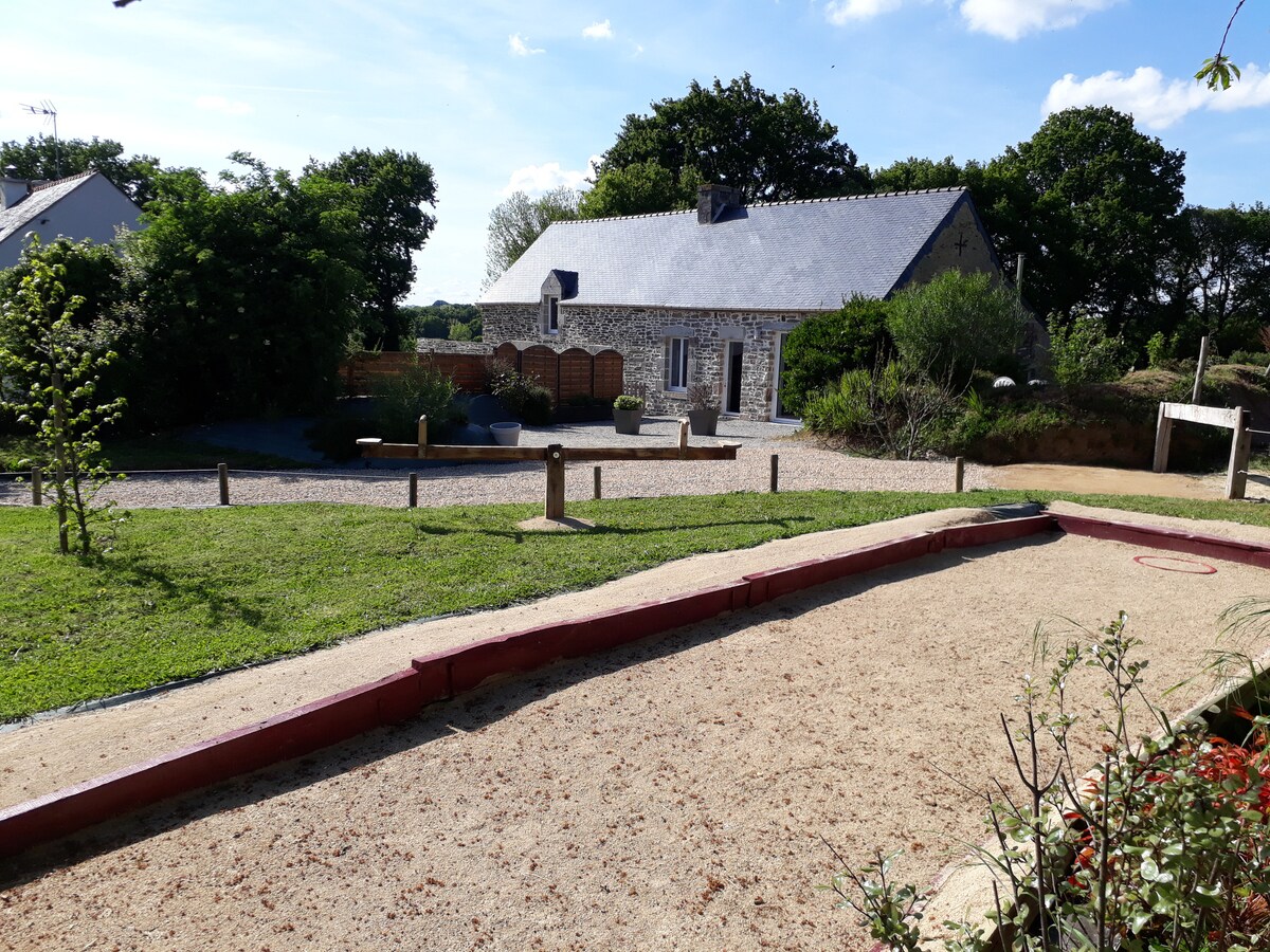A charming stone house is set amidst greenery, featuring a slate roof and large windows. A garden area is visible, complete with a petanque court lined in red. Pathways and landscaping enhance the outdoor space, contributing to a serene countryside atmosphere.