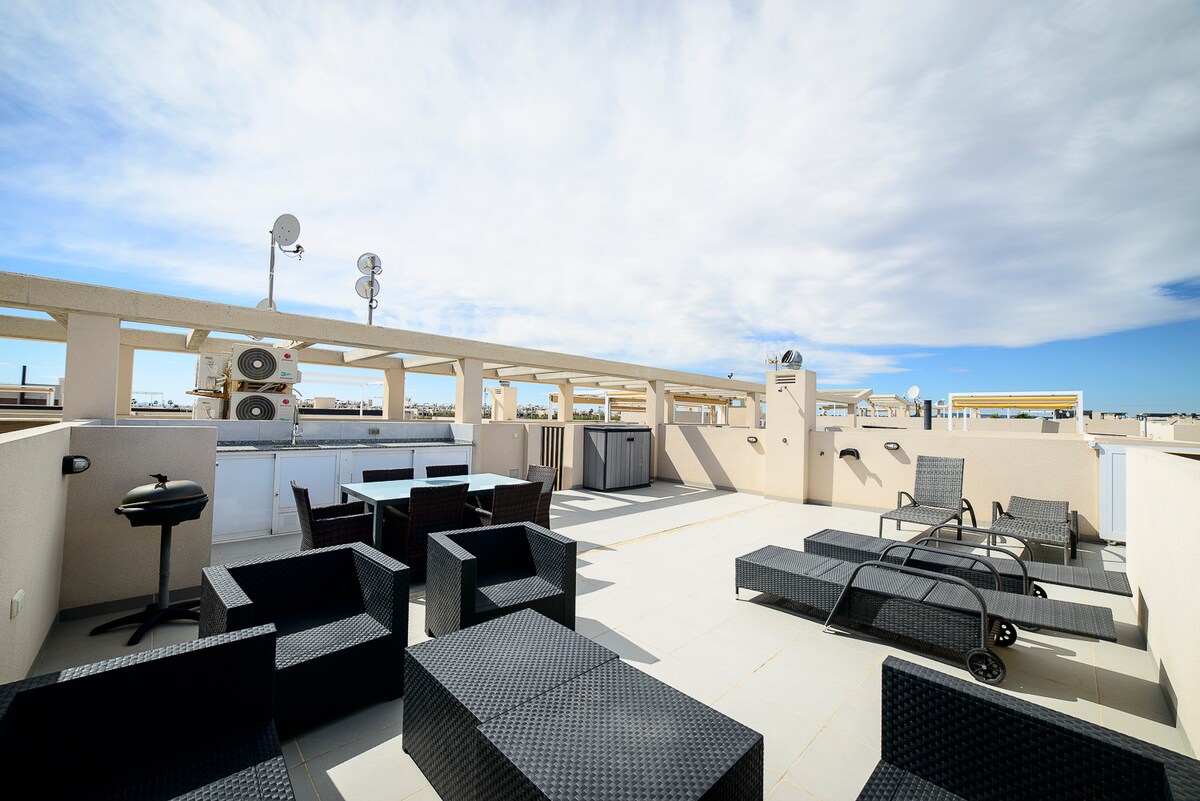 An expansive sun terrace is presented with stylish outdoor furniture, including seating areas and lounge chairs. The space is complemented by clear blue skies and subtle clouds above, creating an inviting outdoor environment for relaxation.
