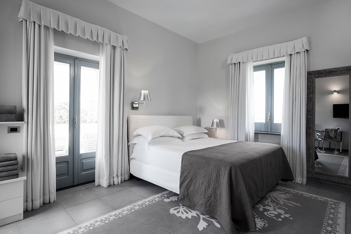 A spacious bedroom features a large bed with neutral bedding, complemented by a decorative gray throw. Two sets of doors lead to a balcony, framed by light curtains. A tall mirror enhances the space, and a cozy seating area can be seen in the background.