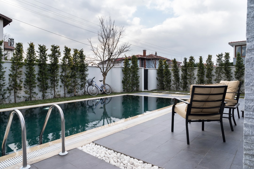 A serene outdoor space features a heated swimming pool surrounded by lush greenery. Two comfortable chairs are positioned nearby, providing a spot to relax. A bicycle is parked beside the pool, adding a touch of casual outdoor living.