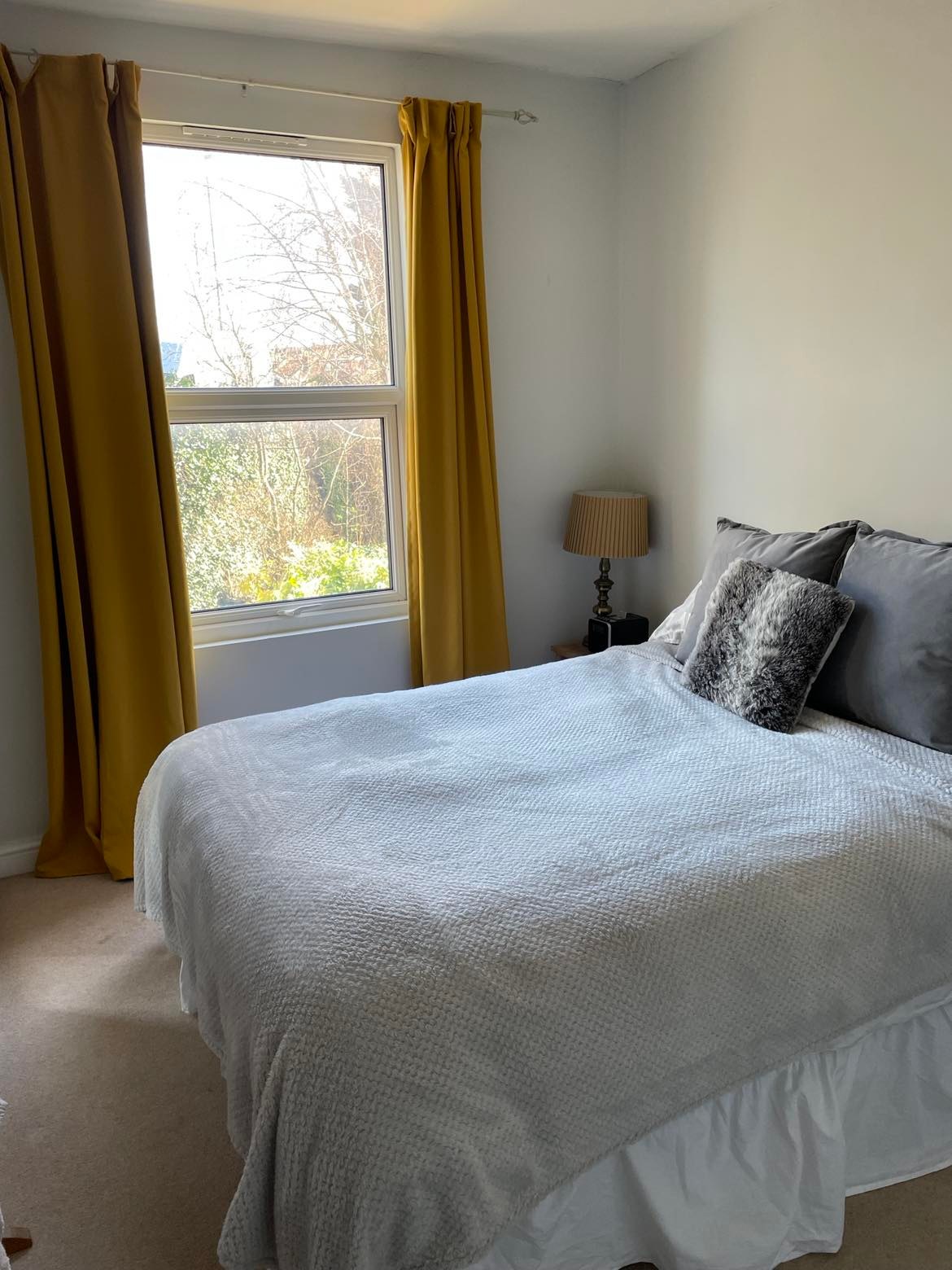 A well-lit bedroom features a spacious bed covered with a light grey quilt. Yellow curtains frame the window, allowing natural light to brighten the room. A small bedside table with a lamp adds functionality to the space, complemented by a decorative pillow on the bed.