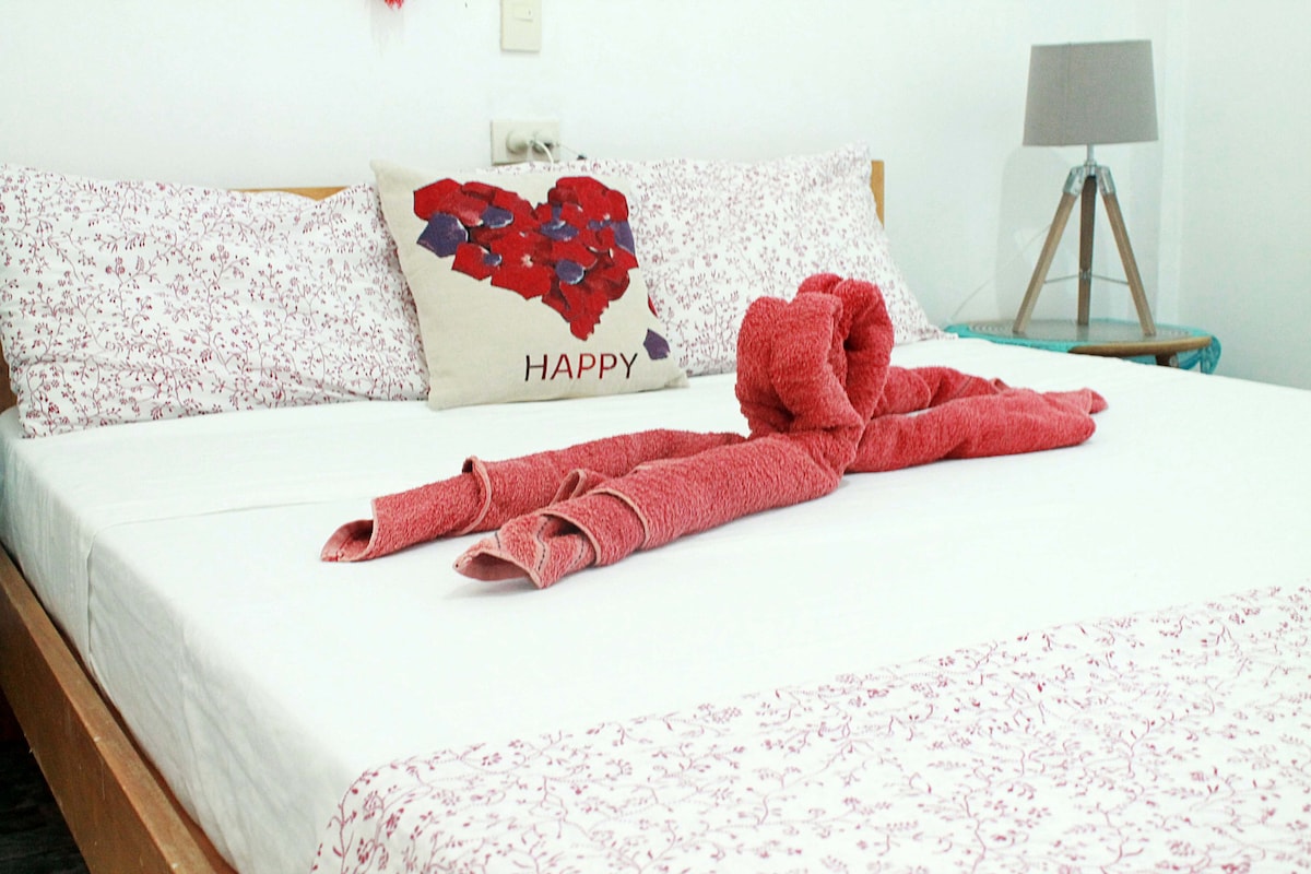 A spacious bed features a minimalistic design with light bedding adorned with a floral pattern. Coordinating pillows sit against the headboard, and a decorative pillow with a heart design adds a touch of charm. A bedside lamp and a small table complement the inviting setup.
