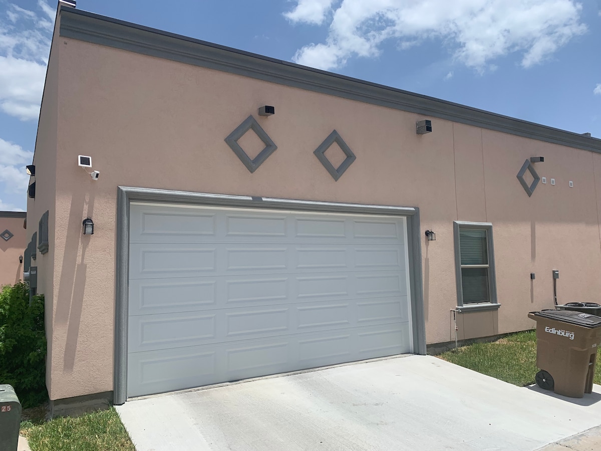 The exterior of a modern townhome is displayed, featuring a light-colored facade with decorative diamond motifs. A spacious two-car garage is present, accompanied by a window on the side. The surrounding area includes well-maintained grass, and the sky is characterized by a few clouds.