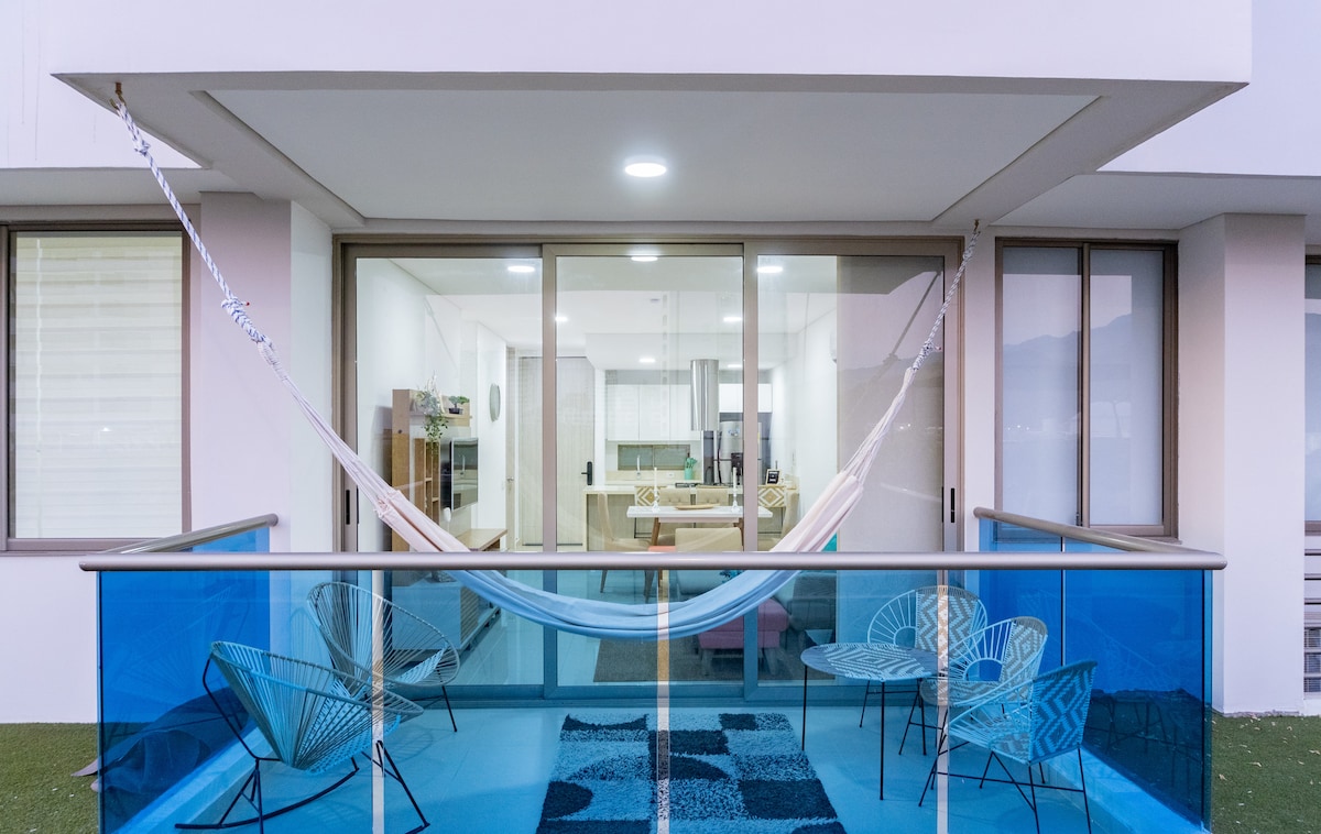 An outdoor terrace features a suspended hammock and four stylish chairs arranged around a patterned rug. Large glass doors provide a view into the interior living space, allowing natural light to fill the area.