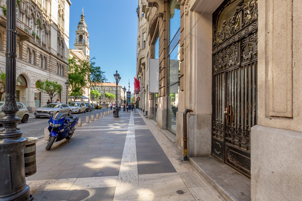 A street view captures the elegant building façade, with intricate wrought-iron details on the entrance gate. Sunlight streams down the bustling sidewalk, where trees line the path. Nearby, a blue motorcycle is parked and a church tower is visible in the distance.