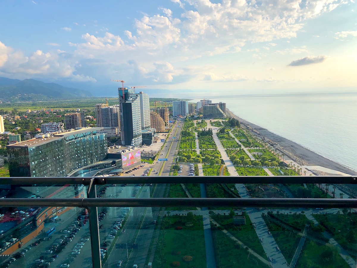 A panoramic view captures the coastline along the beach, featuring a landscaped park and buildings lining the shore. The horizon showcases a mix of mountains and sky as natural light brightens the scene, while modern architecture dominates the foreground.