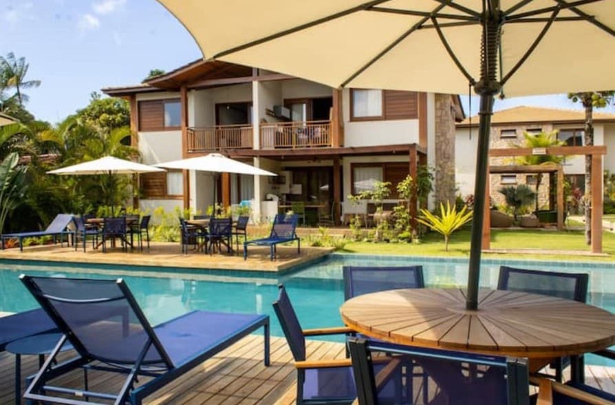 A spacious outdoor area surrounds a sparkling pool, with comfortable lounge chairs arranged beside it. Table seating is provided under a large umbrella. The backdrop features a two-story building with balconies and well-maintained gardens, enhancing the inviting atmosphere.