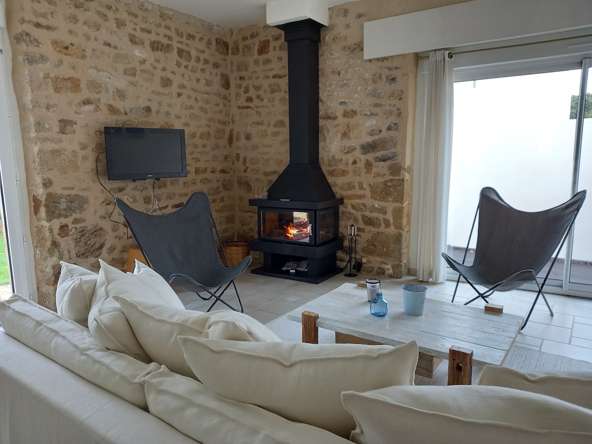 A living area is displayed with a stone wall and a modern wood-burning stove. Two sleek armchairs are positioned near the fire, while a soft sofa with light-colored cushions invites relaxation. Natural light pours in through the nearby glass doors, enhancing the spacious feel.