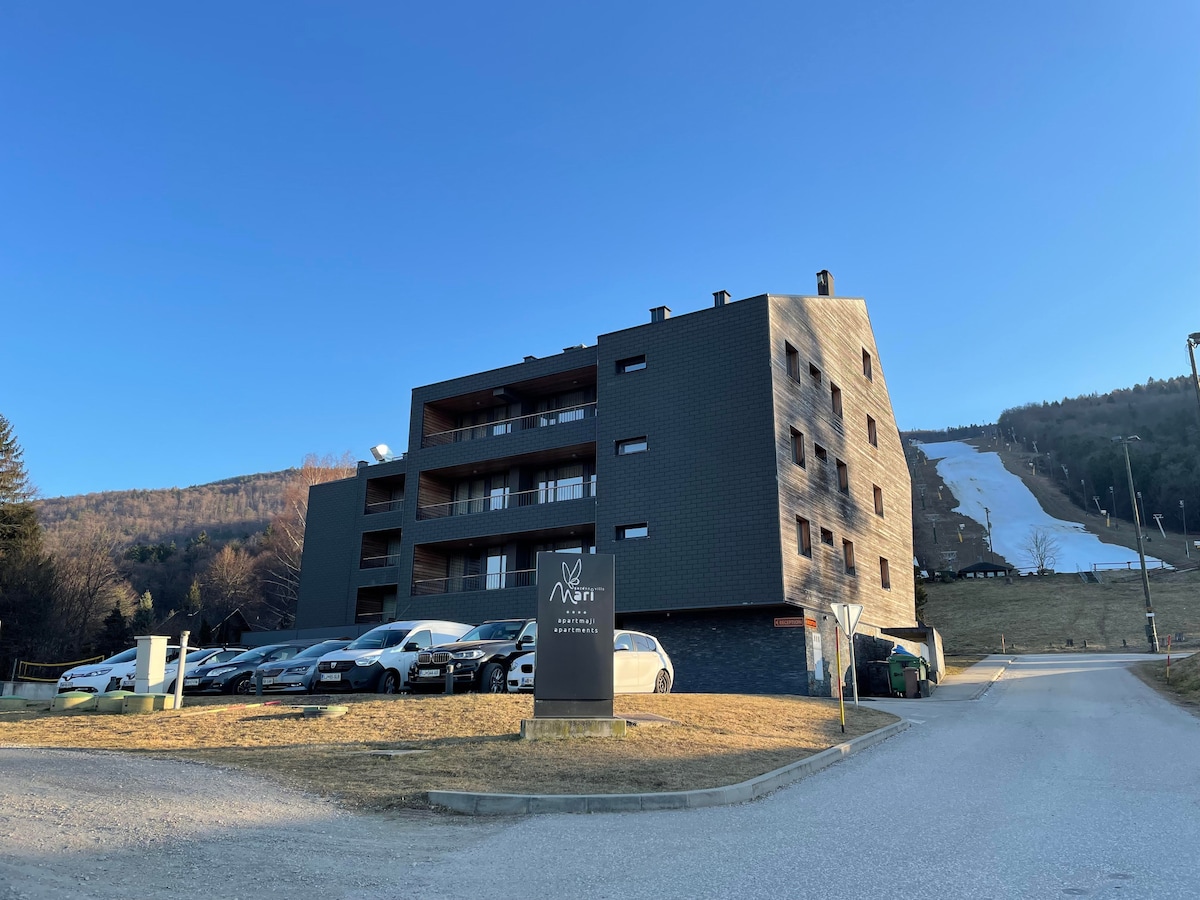 The exterior of the duplex apartment building is presented, featuring a modern design with dark cladding and multiple balconies. A paved area is visible in front, accommodating several parked cars. In the background, the snowy slopes of Mariborsko Pohorje are noticeable.