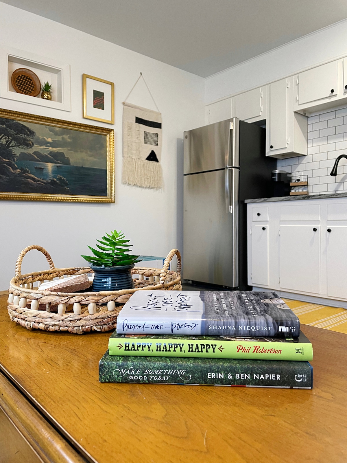 A wooden table displays a stacked arrangement of four books, topped with a small potted plant in a woven basket. In the background, a wall-mounted artwork and a framed landscape painting add detail to the bright kitchen space, with a silver refrigerator visible.