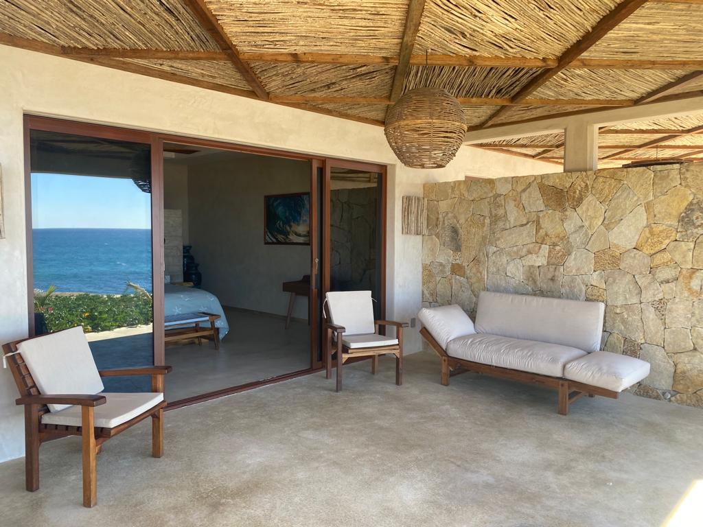 A shaded outdoor seating area features two wooden chairs and a spacious lounge sofa, framed by natural stone walls. The backdrop presents a view of the ocean through large glass doors that open to an indoor space, allowing for a seamless connection with the serene surroundings.