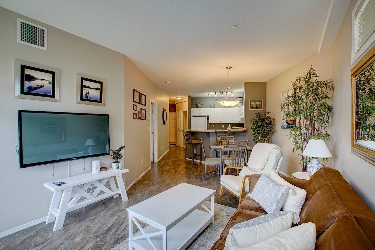 An inviting living area is highlighted by a large flat-screen TV mounted on the wall. Comfortable seating is provided by a white sofa and brown armchair, with a coffee table in the center. The open layout connects to a dining space and kitchen, enriched by decorative plants.