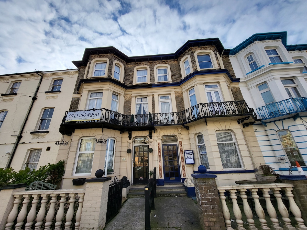 The exterior of the Collingwood guest house is displayed, featuring a multi-story building with a mix of architectural styles. Balconies adorn the upper levels, and the entrance is framed by decorative pillars. A clear sky enhances the welcoming appearance of the property.