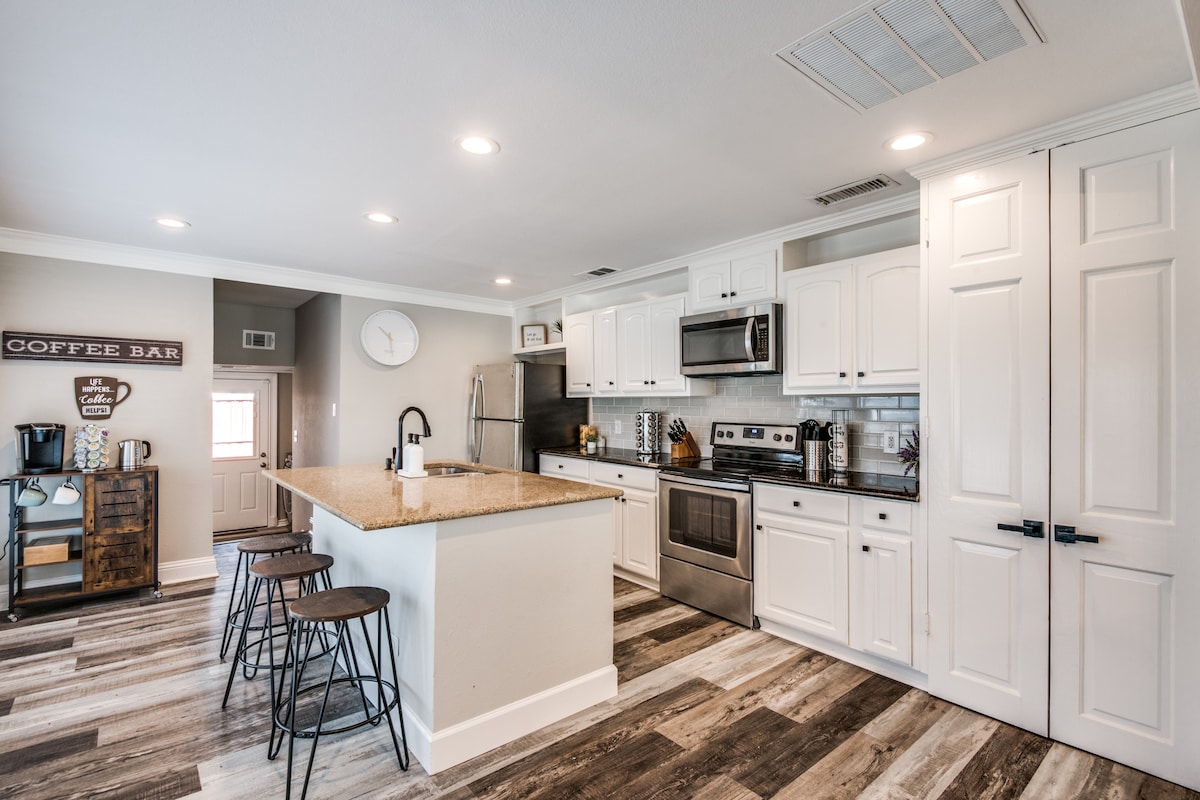 A spacious kitchen is equipped with modern appliances and white cabinetry. A central island with a granite countertop accommodates three stools, while recessed lighting enhances the area. A coffee bar is visible against a wall, with a clock and other decorative items completing the setup.