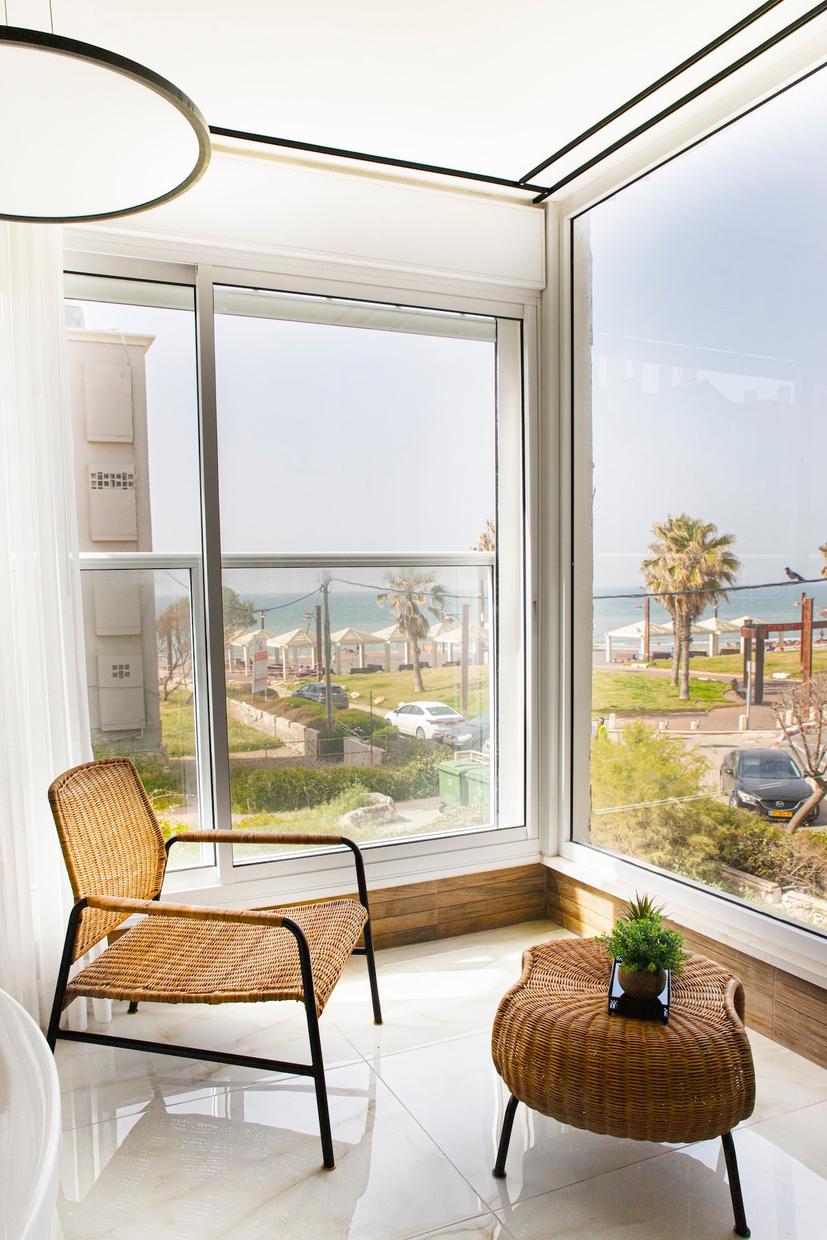 A sunlit corner features two seating options: a rattan chair and a round coffee table positioned by the expansive glass window. The view reveals palm trees, a beach promenade, and the shimmering sea in the distance.