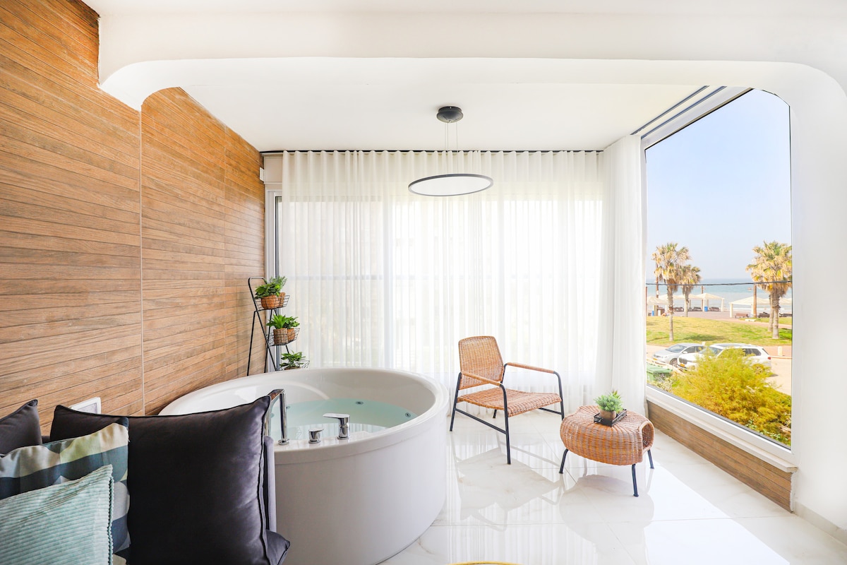 A bright and airy space features a jacuzzi next to large glass windows. The room is complemented by a cozy seating area with two stylish chairs and a small table. Natural light floods the area, providing a view of palm trees and the sea.