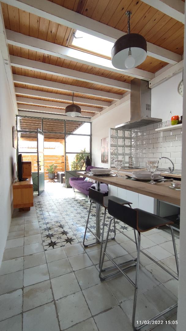 A modern kitchen features a sleek countertop with bar seating, enhancing the open layout. Natural light fills the space through large glass doors, which provide access to a private outdoor area. The floor showcases a distinctive tile pattern, complementing the wooden beams overhead.