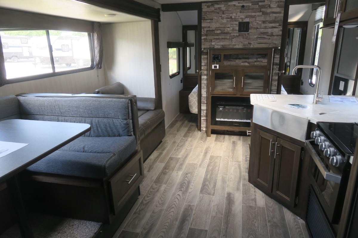 The spacious interior of the camper features a cozy seating area with a gray couch and a dining table. Natural light enters through large windows, enhancing the open layout that includes a kitchen with a refrigerator and stove. A stone accent wall adds a touch of warmth.