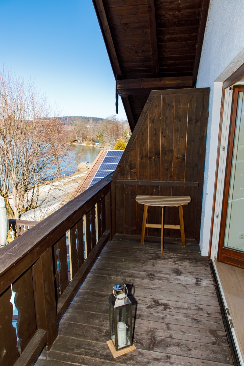 A wooden balcony area features a small, round table and a lantern. The view reveals a serene lakeside scene, enhanced by the clarity of the sky. Natural wood elements and minimalist decor create a simple and peaceful outdoor space.