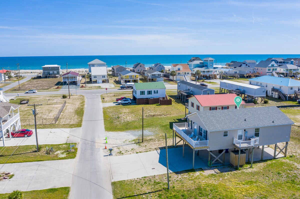An aerial view reveals a coastal home positioned just across the street from the ocean. The surrounding neighborhood features various residences and a clear blue sky, highlighting the proximity to the beach.