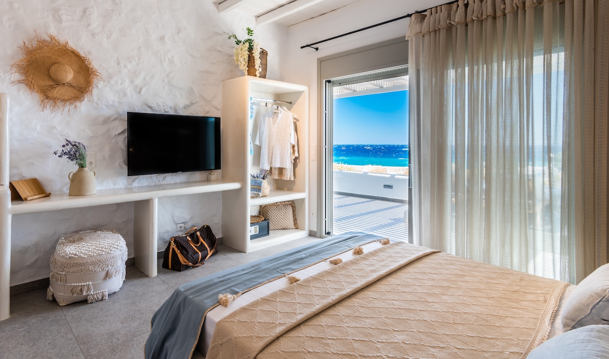 A well-lit bedroom features a queen bed adorned with a light-colored coverlet. A flat-screen TV is mounted on a white wall unit alongside a storage area. Large glass doors lead to a terrace that overlooks the sea, with sheer curtains gently diffusing the light.