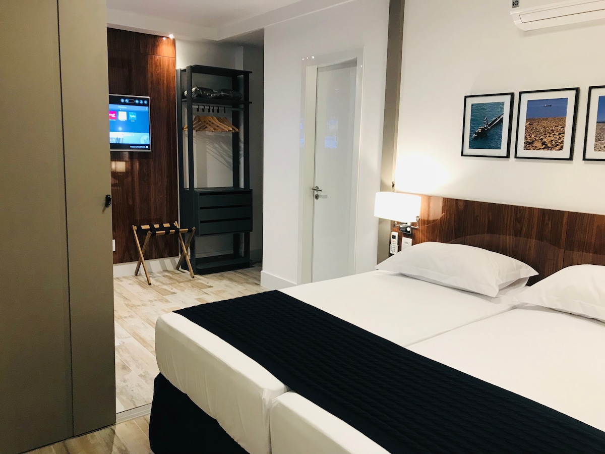 A cozy bedroom features a spacious bed dressed in dark bedding, complemented by light walls and a wooden accent wall. Framed images adorn the wall above the bed. A television is mounted on a nearby wall, and a clothing rack is positioned next to a wardrobe.