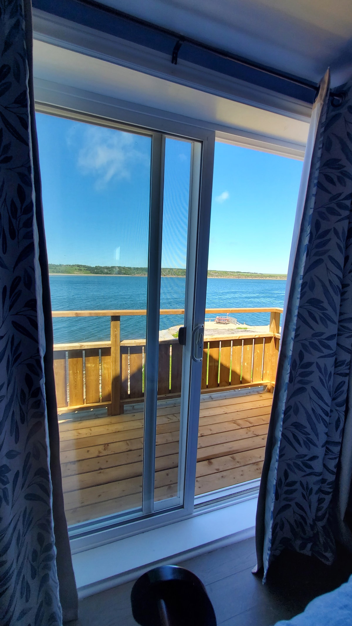 A sliding glass door offers a view of the water, framed by patterned curtains. The spacious deck outside invites relaxation, with wooden flooring and a railing overlooking the serene waters. Bright blue skies complement the tranquil waterfront scene.