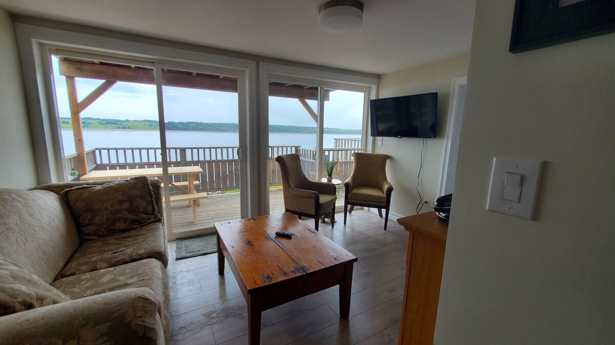 A cozy living area features a comfortable sofa and two armchairs, with a wooden coffee table at the center. Large sliding glass doors provide scenic views of the water and access to the balcony. A television is mounted on the wall opposite the seating.