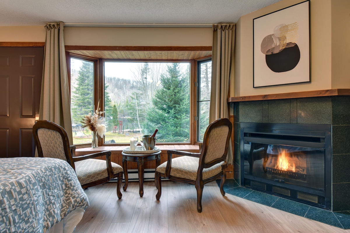 A cozy corner features two elegant chairs around a small table, positioned by large windows offering views of surrounding trees. A fireplace is visible, radiating warmth, while soft natural light filters in from the outside.