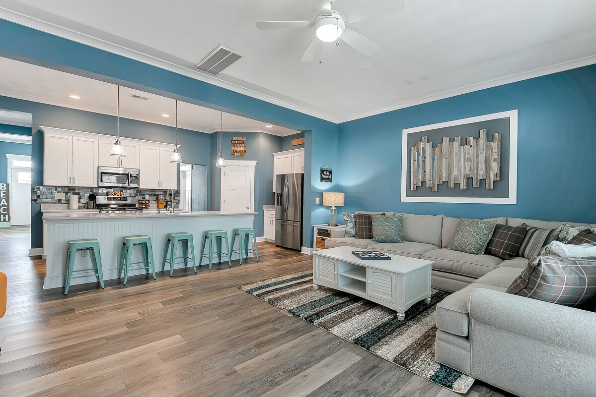 The spacious living room features a light gray sectional sofa adorned with patterned cushions. A wooden coffee table sits atop a soft area rug with varying tones. The open kitchen area is visible in the background, complemented by three green bar stools near a breakfast bar.