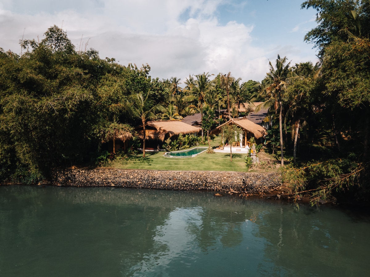 The retreat is set along the Balian River, surrounded by lush greenery and tropical palm trees. A serene swimming pool is visible amidst the landscaped garden, complemented by traditional thatched-roof bungalows, creating a harmonious connection with the natural environment.