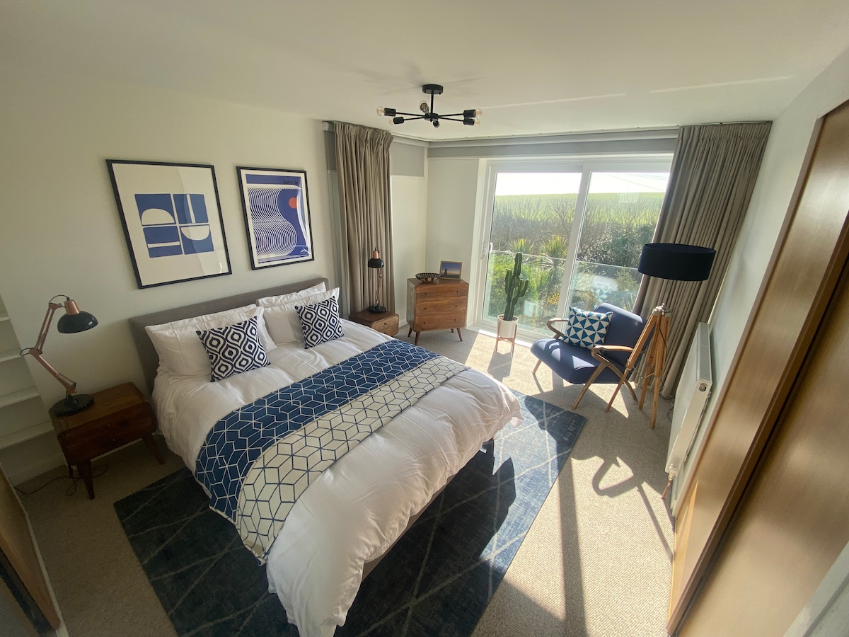 A well-appointed bedroom is presented with a spacious bed adorned in white linens and a blue patterned throw. Framed artwork is displayed above the bed. Natural light filters in through large glass doors, leading to a serene outdoor view. A comfortable chair and stylish lamp are also featured.