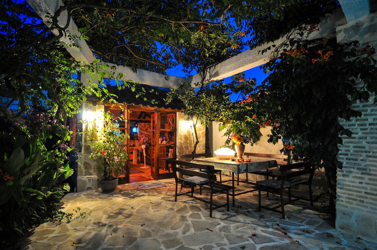 An outdoor patio area is surrounded by lush greenery and softly illuminated by warm lighting. A dining table with several chairs is set for enjoying meals, while the entrance to a cozy indoor space is visible in the background, enhancing the welcoming ambiance.