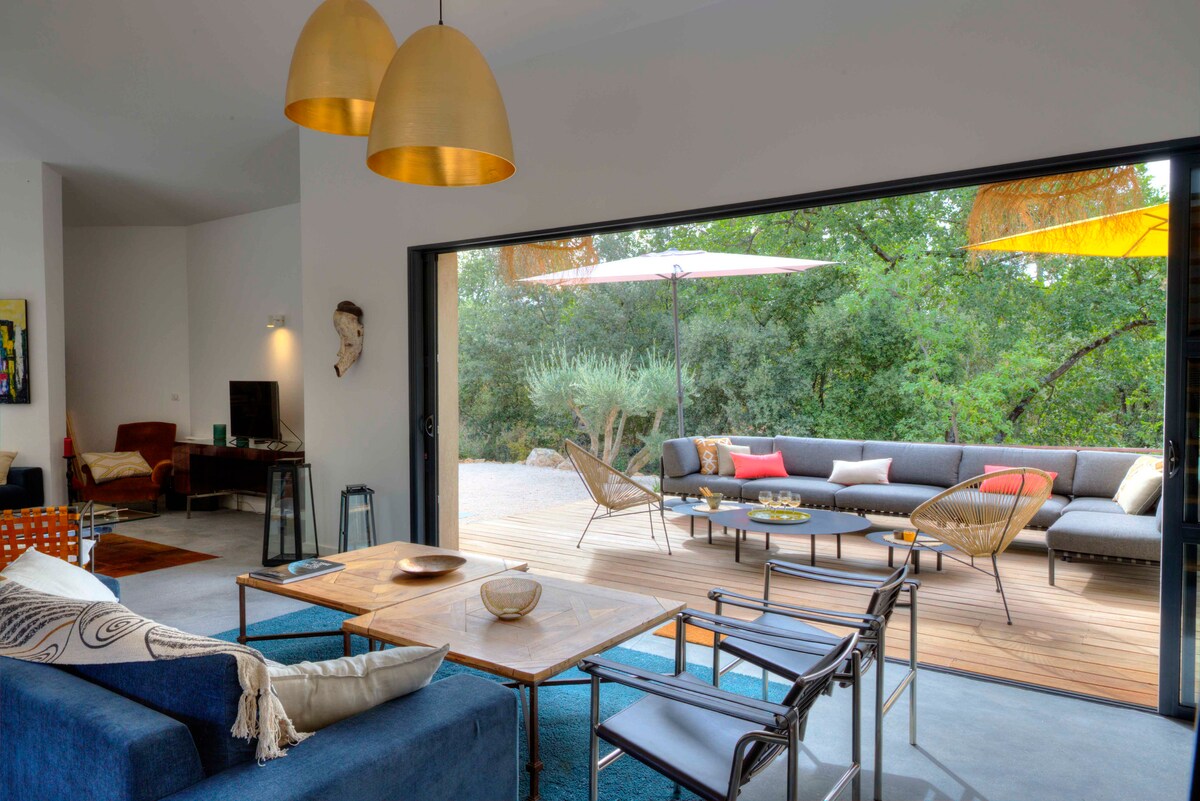 A spacious living area is showcased with contemporary furniture, including a wooden table and comfortable seating. Wide glass doors open to an outdoor terrace featuring lounge seating and umbrellas, surrounded by lush greenery. Warm lighting fixtures enhance the inviting atmosphere.