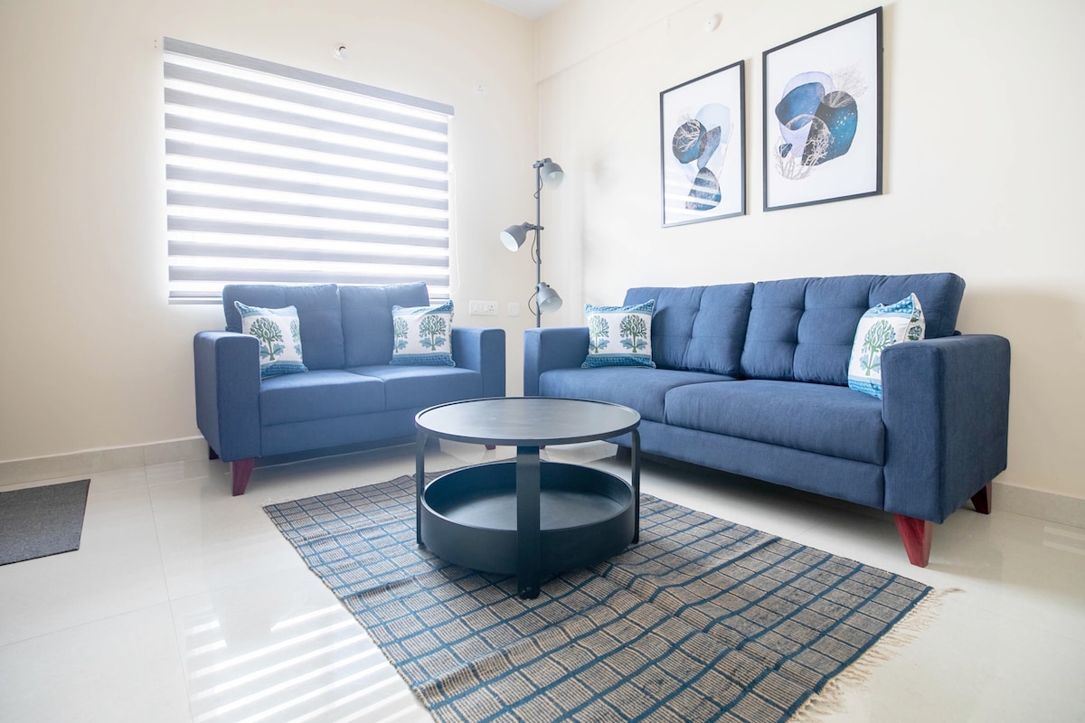 A bright living area features two blue sofas adorned with decorative cushions. A round coffee table is positioned centrally on a textured rug. Artwork is displayed on the wall, and natural light filters through a horizontal striped window treatment.