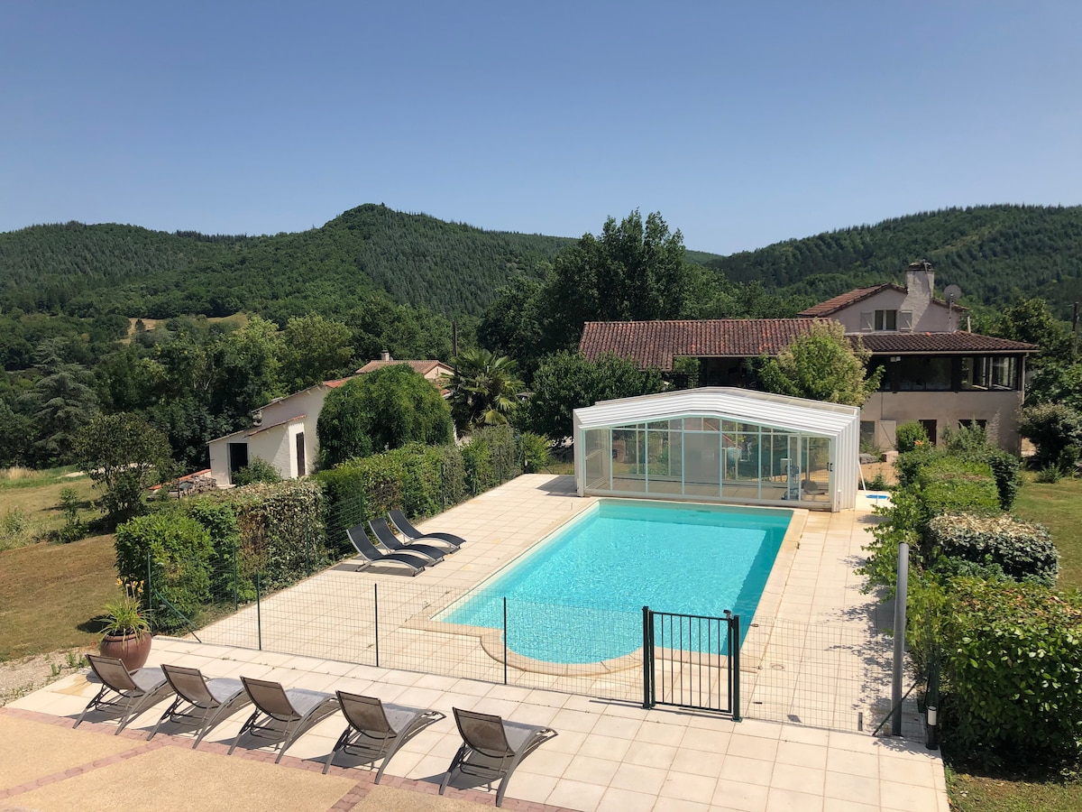 A shared swimming pool is enclosed under a protective structure, surrounded by a spacious patio area. Sun loungers are arranged around the pool, with lush greenery and rolling hills visible in the background, creating a serene environment.