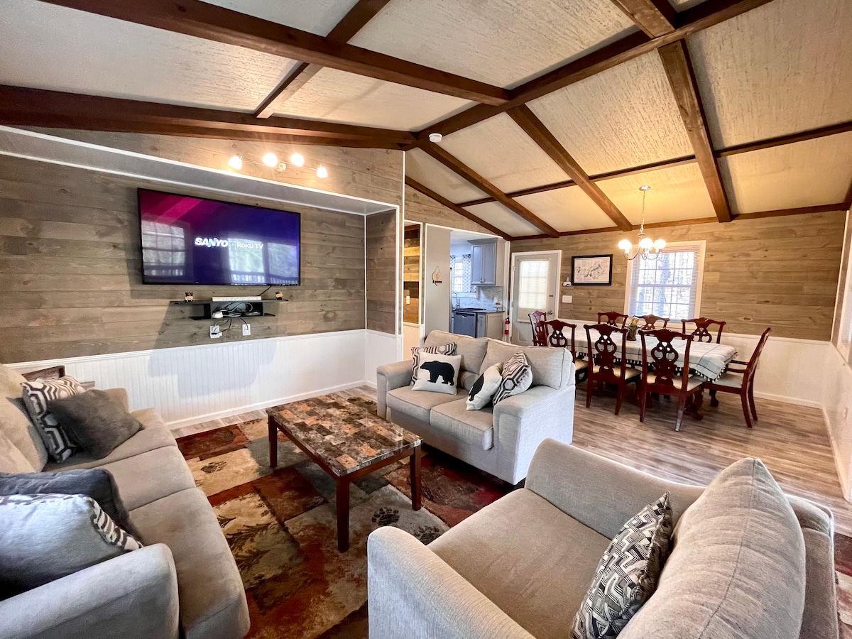 A cozy living area features two light-colored sofas arranged around a rustic coffee table. A wall-mounted television is visible above a white panel, with natural light streaming through nearby windows. The adjoining dining area showcases a wooden table set for six beneath a chandelier.