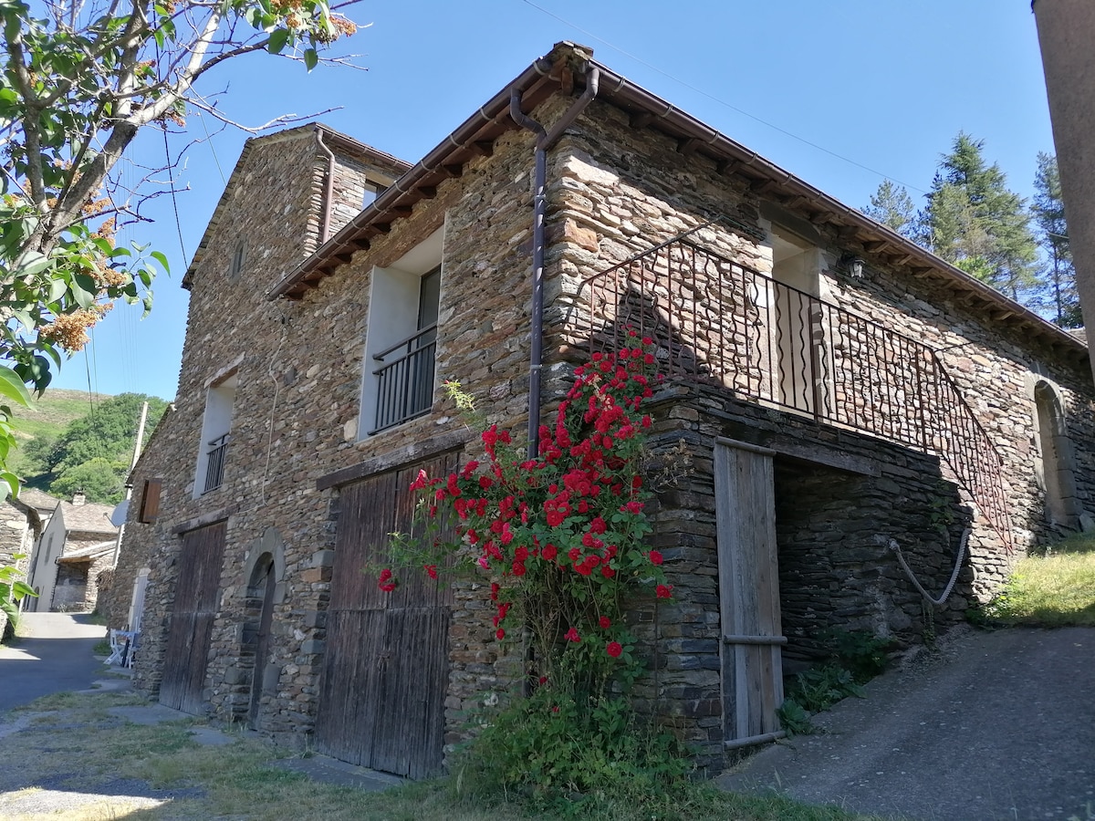The exterior of a renovated stone barn is shown, featuring a rustic facade with wooden accents. A covered balcony extends from the upper level, while vibrant red flowers climb the stone wall. The surrounding area includes greenery and hints of the serene countryside.
