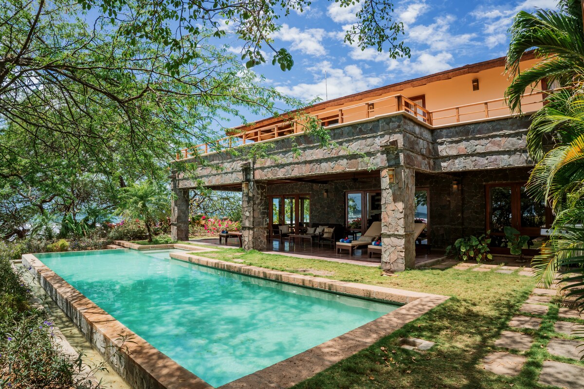 A spacious outdoor area features a lush garden and a clear swimming pool, surrounded by stone walls. Comfortable lounge chairs are positioned nearby, and a view of the ocean can be seen through the greenery. The house above has a balcony overlooking the pool.
