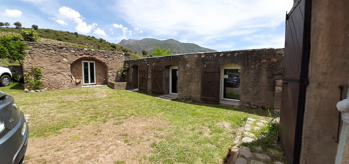 A charming outdoor area is showcased, featuring a grassy lawn bordered by stone pathways. The rustic stone structure displays two entrance doors, while natural scenery and gentle slopes are visible in the background. A car is parked in a sheltered spot, enhancing accessibility.