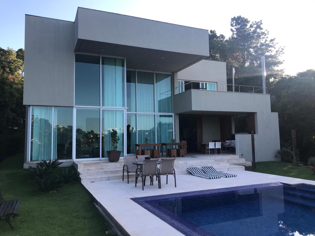 The modern structure features large glass windows, allowing natural light to fill the interior spaces. An outdoor area includes a swimming pool, complemented by lounge chairs and a dining table. Lush greenery surrounds the property, enhancing the serene mountain setting.