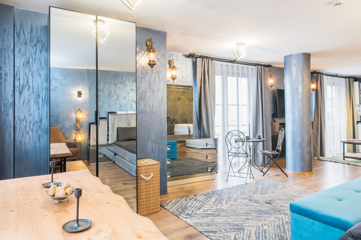 A spacious living area features modern furnishings, including a large blue sofa and a wooden dining table. Natural light filters through sheer curtains, complementing the stylish decor. A mirrored wall enhances the sense of space. Two chairs and a hot tub are visible in this inviting setting.