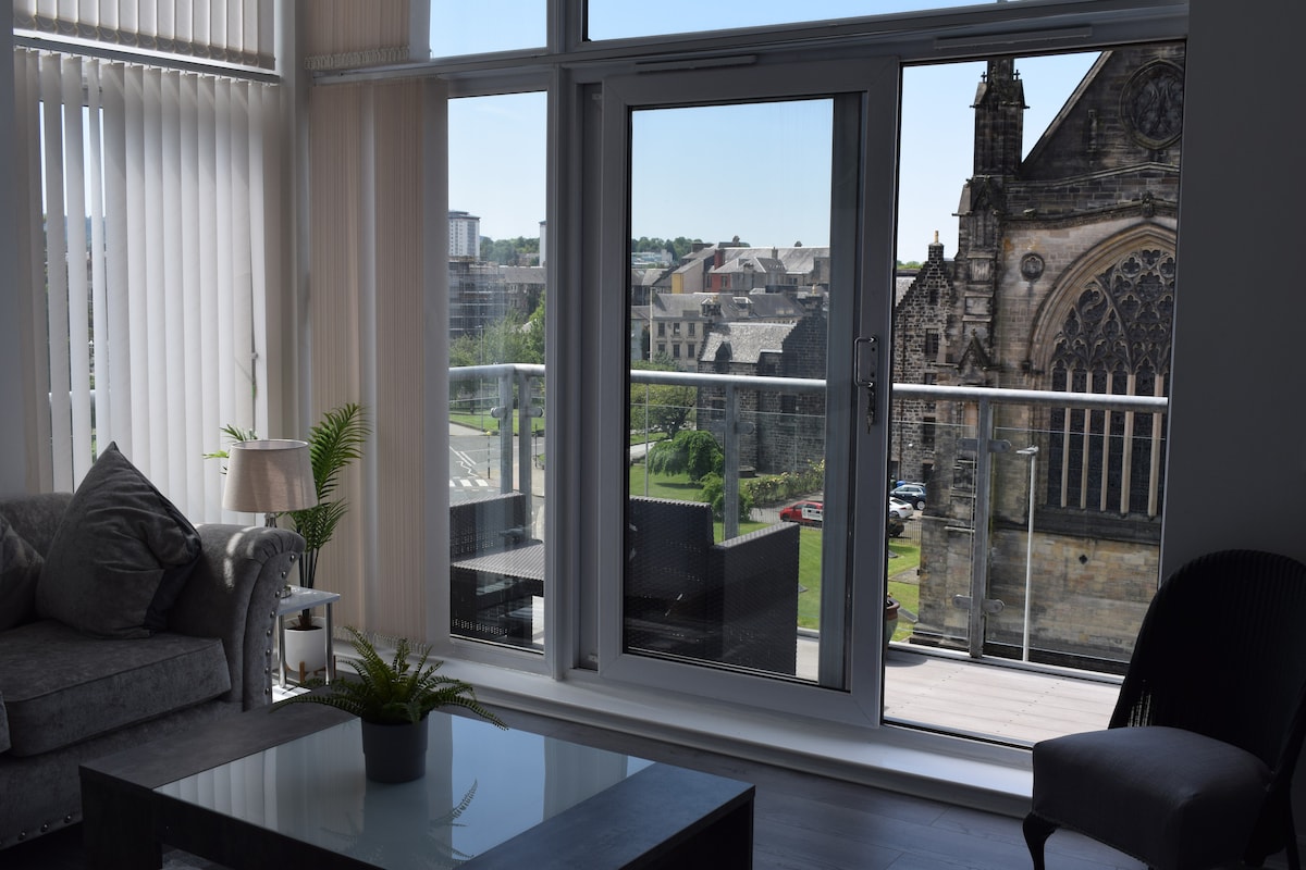 A spacious living area opens onto a balcony, showcasing expansive views of the historic Paisley Abbey. Sunlight filters through large glass doors, illuminating a comfortable seating arrangement and a coffee table. Nearby, a small plant adds a touch of greenery to the space.