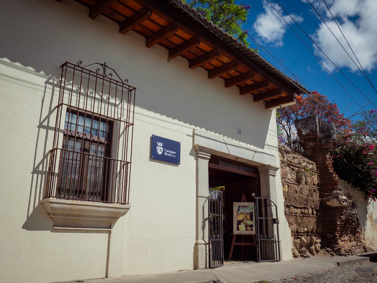 The entrance of Cacique Real Hotel is framed by a classic façade with wooden beams and ornate metalwork on the windows. A welcoming sign is positioned next to the doorway, complemented by greenery. Bright blue skies and scattered clouds provide a vibrant backdrop.