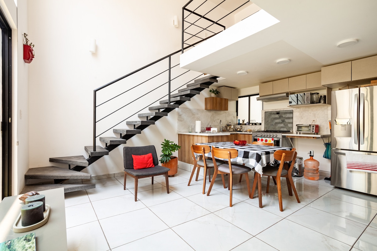 A modern kitchen and dining area are featured, with a large dining table surrounded by wooden chairs. A black sofa is positioned nearby, and a sleek staircase leads to the upper level. Natural light fills the space through large windows, highlighting neutral tones and green plants.