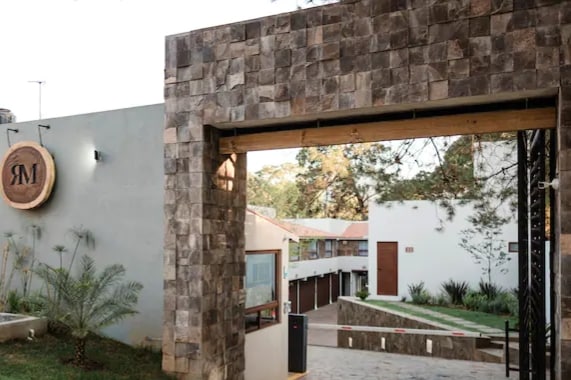 A modern entrance is framed by a stone archway, leading to a landscaped path lined with greenery. The property features a logo sign and a glimpse of the building's clean, white walls in the background.