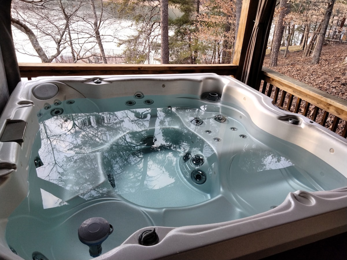 A spacious hot tub is situated on a wooden deck, providing a view of the tranquil waters beyond. The water is clear and inviting, reflecting the surrounding trees and natural landscape.