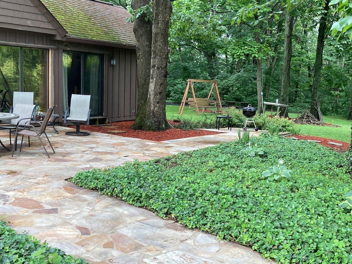 A stone patio area is surrounded by lush greenery, featuring outdoor seating with two chairs and a small table. A swing set and a charcoal grill are positioned nearby, complemented by red mulch that enhances the natural setting.