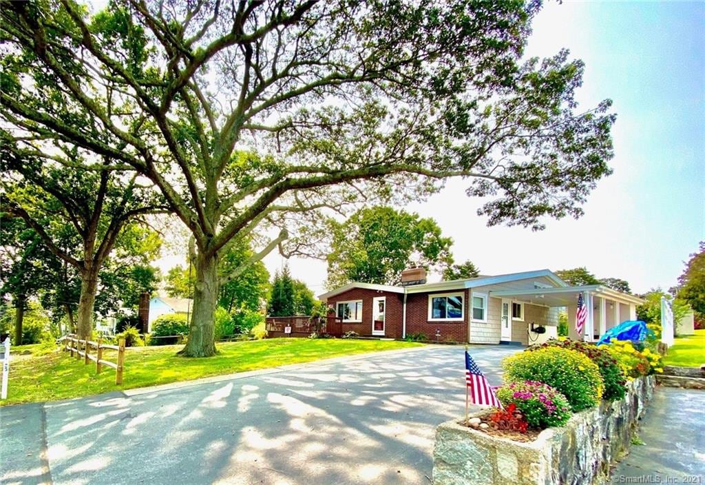 A spacious driveway leads up to a split-level house set amongst large trees. A well-maintained yard is visible, with areas for relaxation. American flags are displayed, and the front entrance is framed by greenery, providing a welcoming view to the property.